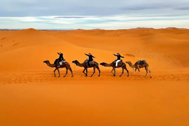 Viajeros en fila sobre dromedarios atravesando las dunas de Erg Chebbi, en Merzouga, con el sol cayendo